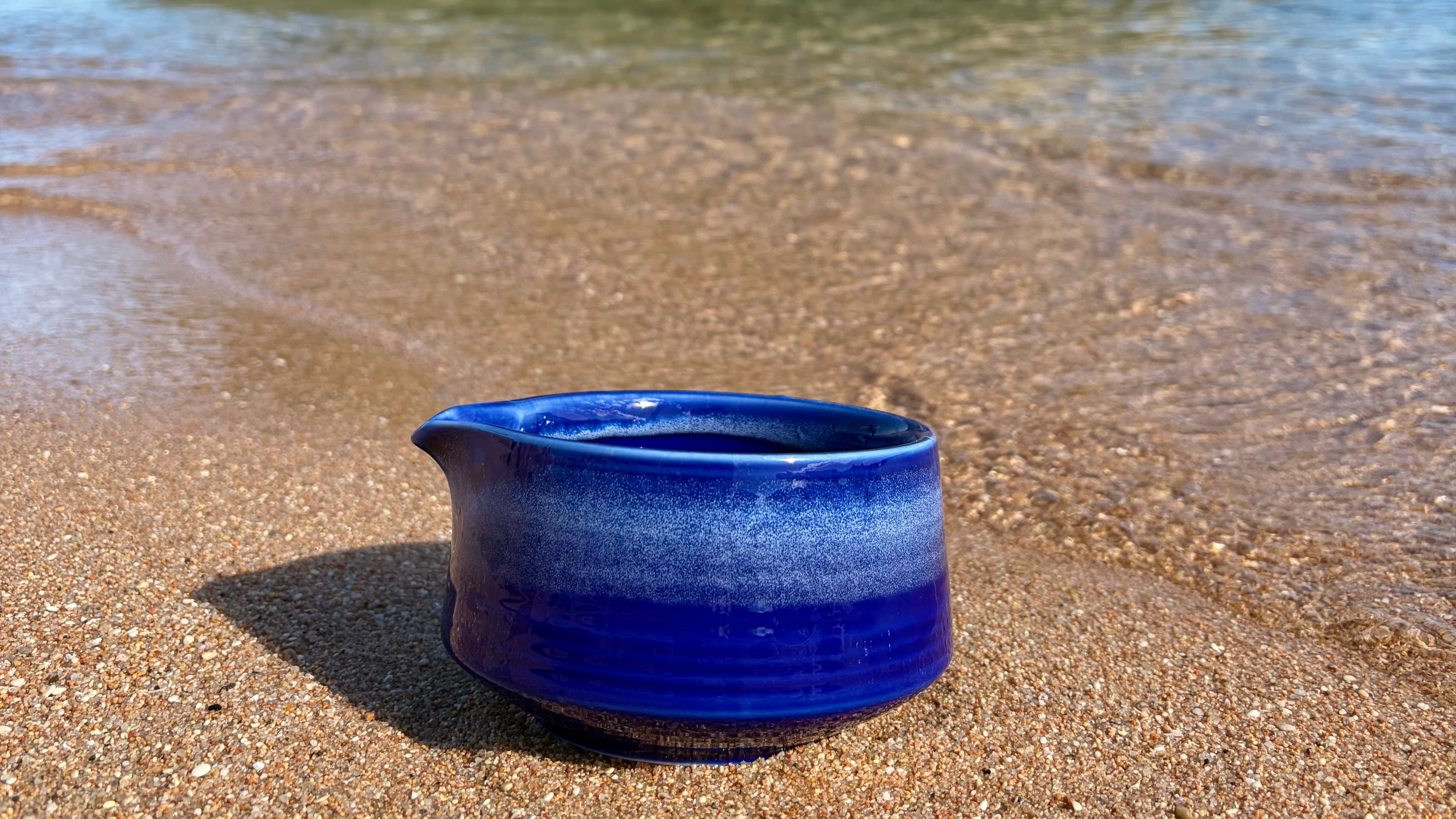 A small glazed ceramic pitcher with a deep cobalt blue and white gradient finish, sitting on wet sand near the shoreline with gentle waves in the background.