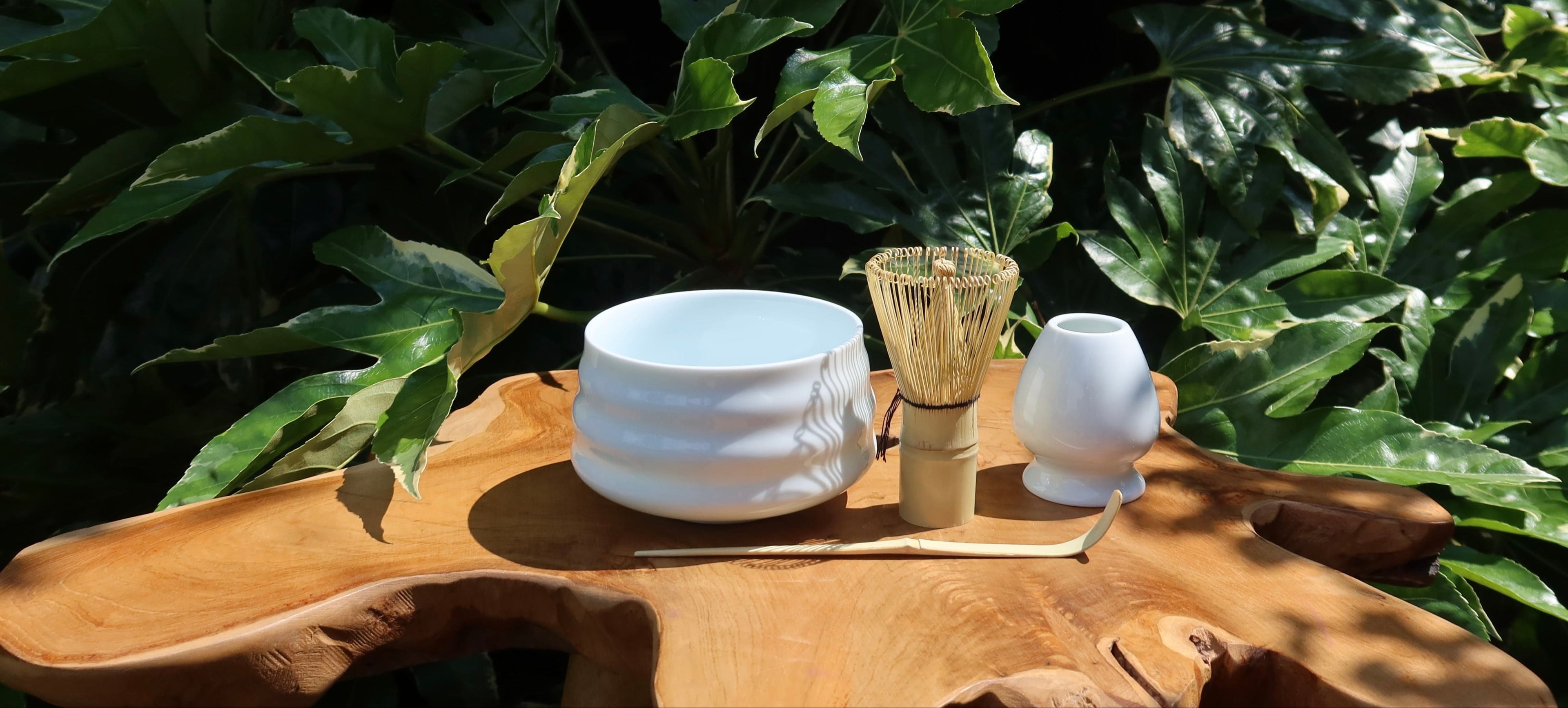 white matcha tea-making set with bamboo whisk on a natural wooden table with green leaves in the background
